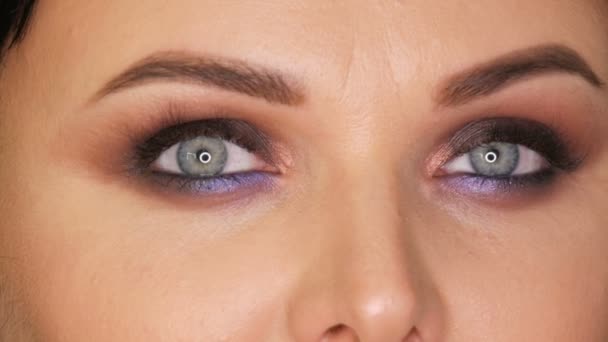 Mujer hermosa joven con ojos azules brillantes y maquillaje de noche ojos ahumados con sombra de ojos azul y púrpura sobre fondo negro de cerca
 - Metraje, vídeo
