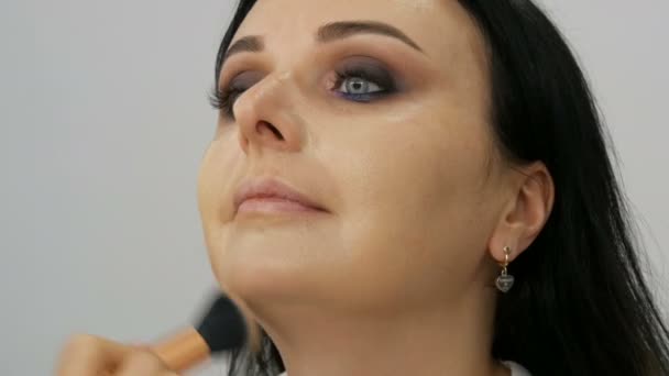 Close up view of a stylist makeup artist applies foundation cream with a special brush on the face of a young beautiful woman with blue eyes - Filmagem, Vídeo