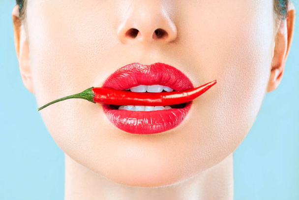 Νεαρή γυναίκα με κόκκινη πιπεριά τσίλι απομονωμένη σε μπλε φόντο. Σέξι γυναικεία χείλη. Καυτό σαγηνευτικό κορίτσι - Φωτογραφία, εικόνα