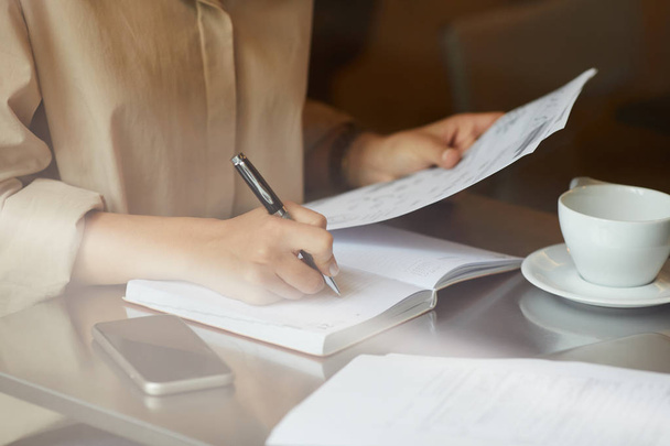 Nahaufnahme einer jungen Geschäftsfrau, die ein Dokument in der Hand hält und Zukunftspläne im Notizblock schreibt, während sie mit einer Tasse Kaffee am Tisch sitzt - Foto, Bild