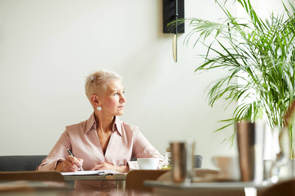 Rijpere zakenvrouw met kort blond haar op zoek weg tijdens het werken aan de tafel op kantoor - Foto, afbeelding