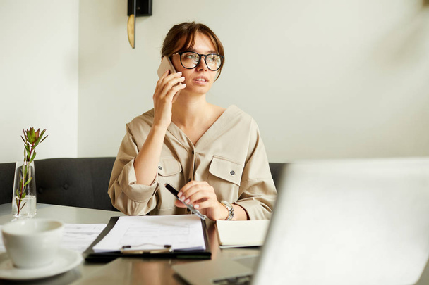 Mladá podnikatelka v brýlích sedí u stolu před notebookem a mluví na mobilním telefonu v kavárně - Fotografie, Obrázek