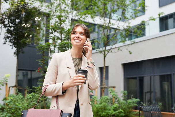 Jonge glimlachende zakenvrouw praten aan de telefoon en het drinken van koffie uit wegwerpbeker tijdens het lopen langs de straat in de stad - Foto, afbeelding