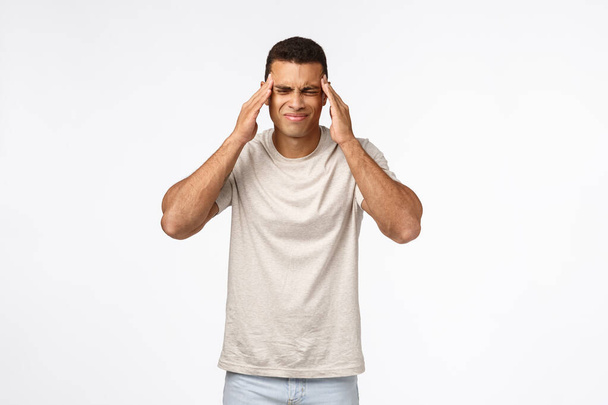 Handsome brazilian guy suffering terrible headache, grimacing from pain, squinting and touching temples, have migraine or ache from hangover, standing white background, feeling not well - Photo, Image