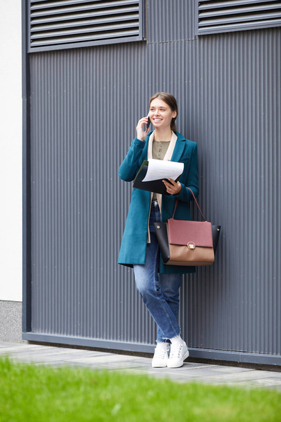 Zarif ceketli, çantalı, duvara yaslanmış ve cep telefonuyla konuşan genç bir kadın. - Fotoğraf, Görsel