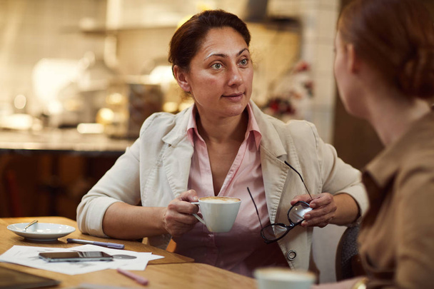 Dojrzała bizneswoman trzyma filiżankę kawy i rozmawia z partnerem przy stole podczas spotkania w kawiarni - Zdjęcie, obraz
