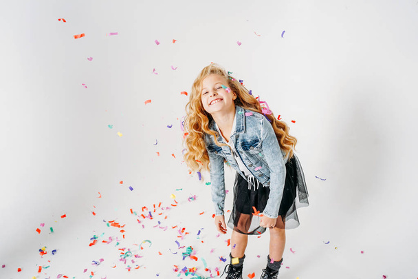 Happy fashionably dressed curly hair tween girl in in denim jacket and black tutu skirt and rough boots on white background with colorful confetti - Foto, afbeelding