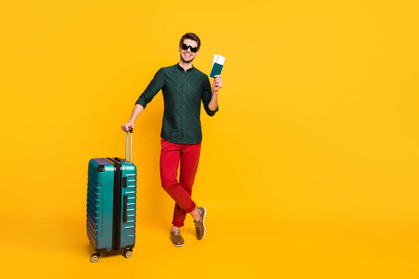Full length photo of positive cheerful brunet hair guy feel content ready to travel voyage hold trolley tickets wear youth outfit red pants trousers sneakers isolated bright color background - Zdjęcie, obraz