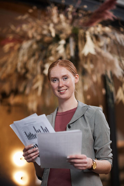 Retrato de una joven mujer de negocios pelirroja sonriendo a la cámara mientras trabaja con documentos en la oficina - Foto, imagen