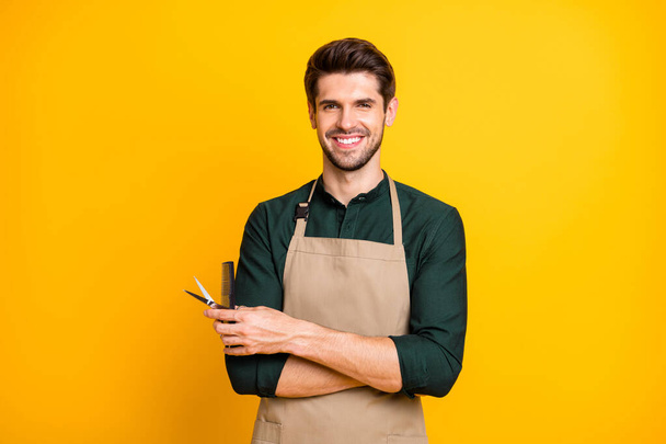Photo of brown haired toothy attractive beaming man holding hair brush and scissors with hands folded professional master in apron isolated over yellow vibrant color background - Foto, immagini