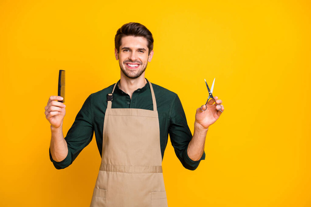 Portrait of positive cheerful guy freelancer ready to make haircut to his clients in modern barbershop hold comb scissors wear stylish trendy green shirt isolated over yellow color background - Fotó, kép