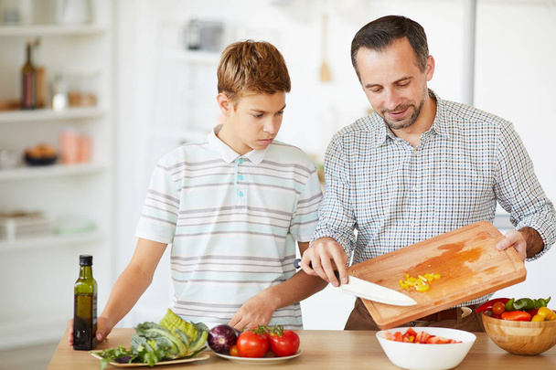 Young father cooking vegetable salad and teaching son to cook in domestic kitchen - 写真・画像