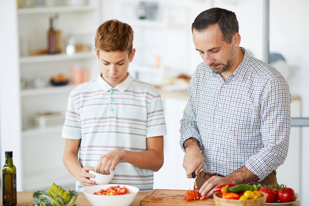 Padre joven cocinando ensalada de verduras y enseñando a su hijo a cocinar en la cocina doméstica
 - Foto, Imagen