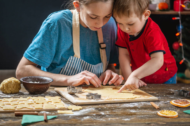 Madre e hijo dan forma a la masa para hornear pan de jengibre casero de vacaciones. Cocina familiar en decoraciones navideñas - Foto, Imagen