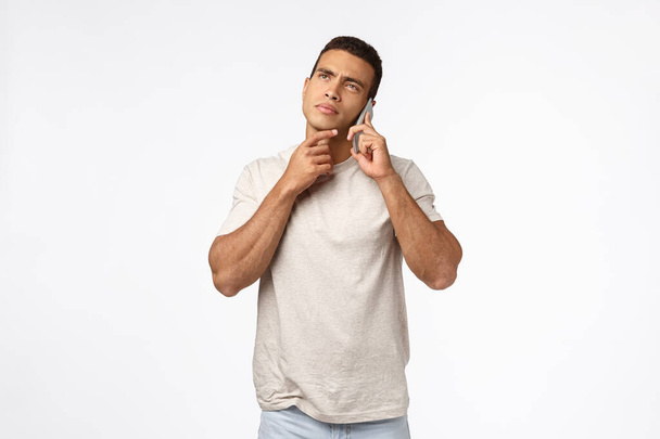 Thoughtful handsome young man in t-shirt, holding smartphone near ear, having conversation, touch chin and frowning, pondering choices, thinking how to answer, making decision, white background - Photo, Image