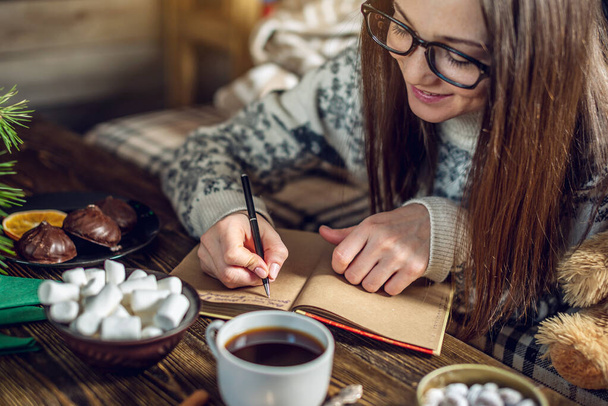 Mulher de camisola está escrevendo uma lista de desejos em um caderno na atmosfera calorosa do Natal. Aconchegante clima de Ano Novo
 - Foto, Imagem
