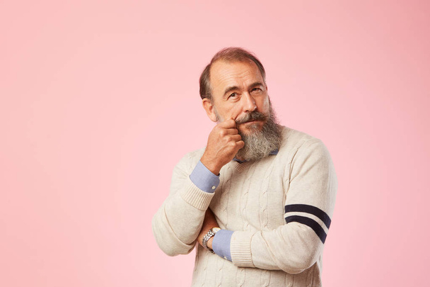 Портрет пожилого человека с седой бородой в белом свитере, задумчиво смотрящего на розовый фон
 - Фото, изображение