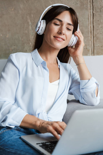 Pihentető nő otthon ül a kanapén a nappaliban zenét hallgatni fejhallgatóval laptoppal. - Fotó, kép