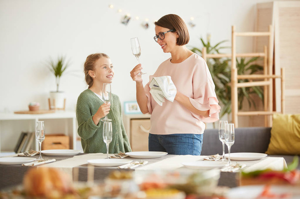 junge Mutter wischt die Weingläser mit Handtuch ab, während Tochter zu Hause am Esstisch steht - Foto, Bild