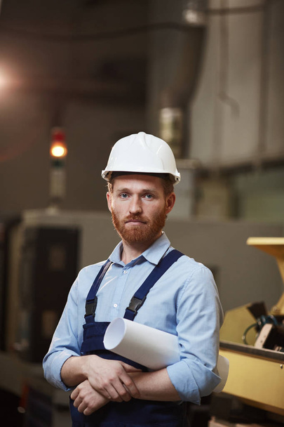 Портрет серьезного бородатого менеджера в комбинезоне и в рабочем шлеме, держащего в руках синий принт и смотрящего в камеру во время работы на заводе - Фото, изображение