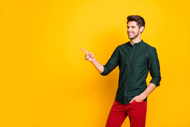 Photo of cheerful positive attractive guy pointing at empty space with hand in trousers pocket smiling toothily isolated vibrant color background - Fotografie, Obrázek
