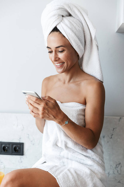 Smiling happy woman in towel after spa procedure shower indoors at home at the kitchen using mobile phone. - 写真・画像