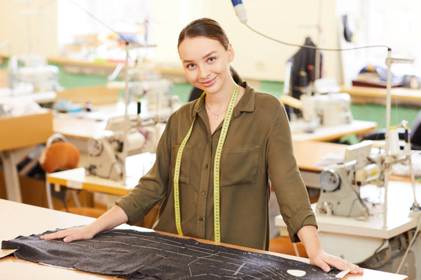 Портрет молодої жінки, що стоїть біля робочого місця з тканиною і дивиться на камеру в майстерні
 - Фото, зображення