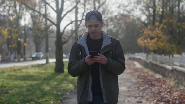 Smart young man walking in  a park and surfing the net in autumn in slow motion      Impressive view of a clever young man in a cap and a jacket walking in a park along a stone parapet and browsing the net on a sunny day in summer in slo-mo - Кадры, видео