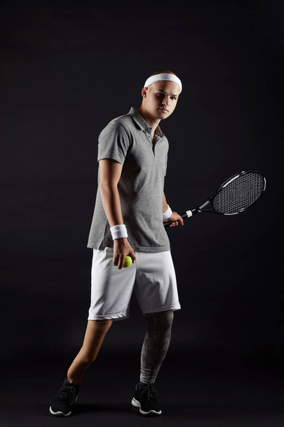 Портрет молодого спортсмена-инвалида в спортивной одежде, держащего ракетку и позирующего перед камерой на черном фоне
  - Фото, изображение