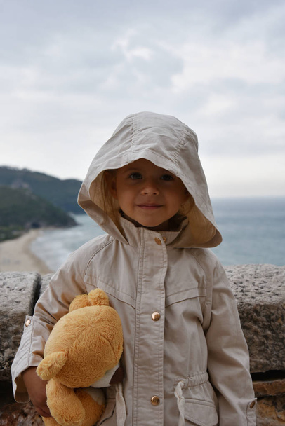 Маленька дівчинка з плюшевим ведмедем носить куртку з капюшоном на бурхливе небо. Концепції дитинства. Екологічні концепції. Концепції клімату
. - Фото, зображення