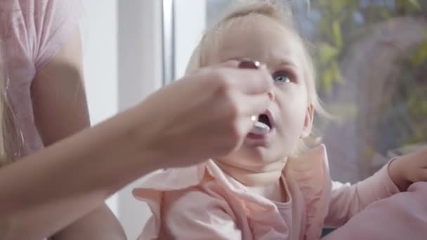 Close-up face of Caucasian little girl eating baby food. Mother feeding her charming child with deep grey eyes. Happy motherhood, joy of maternity. - Video, Çekim