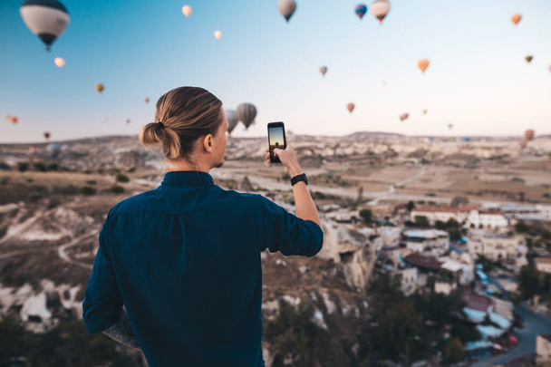 Man taking photo of beautiful landscape and balloons in Cappadocia with mobile camera, sunrise time. - Фото, изображение