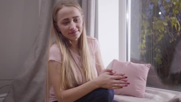 Portrait of young blond Caucasian woman sitting at low windowsill, clapping hands and talking. Pretty mother waiting for her child to come up. Happy parenthood, joy of maternity. - Filmagem, Vídeo