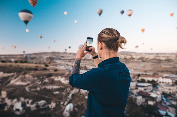 Man taking photo of beautiful landscape and balloons in Cappadocia with mobile camera, sunrise time. - Foto, afbeelding