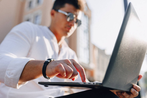 mladý stylový chlap v košili s telefonem a notebookem na lavičce na slunném teplém dni venku, na volné noze - Fotografie, Obrázek