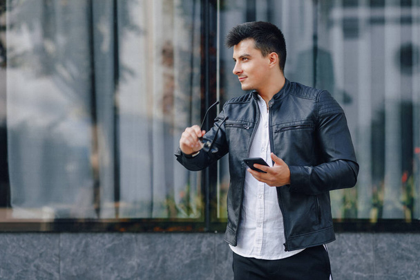 young stylish guy in glasses in black leather jacket with phone on glass background - Photo, image