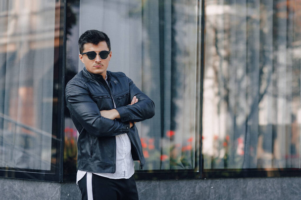 young stylish guy in glasses in black leather jacket on glass background - Zdjęcie, obraz