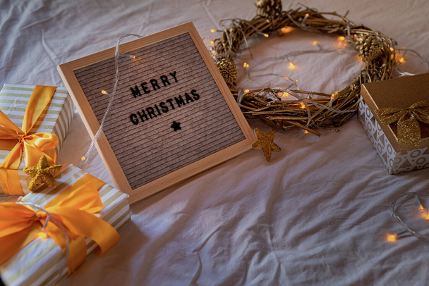 Viltbord "Vrolijk kerstfeest" op het bed versierd met gouden krans, verlichting en geschenkdozen - Foto, afbeelding