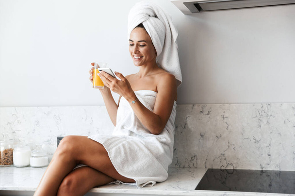 Smiling happy woman in towel after spa procedure shower indoors at home at the kitchen drinking juice using mobile phone. - Foto, immagini