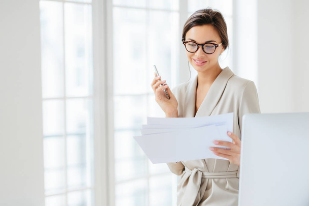 Photo of satisfied young woman with dark hair dressed in business suit, focused in papers, works in office, holds modern cellphone, wears optical glasses for good vision, has pleased expression - Foto, immagini