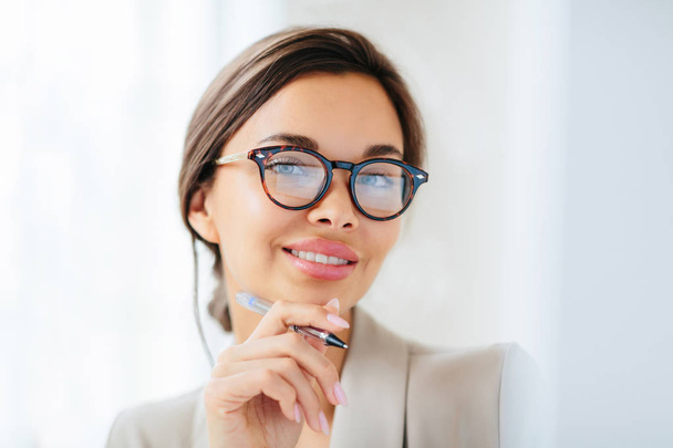 Pleasant looking young female model with makeup and manicure holds pen, touches chin gently, looks directly into camera, dressed in formal clothing, works in office of company, wears optical glasses - Φωτογραφία, εικόνα