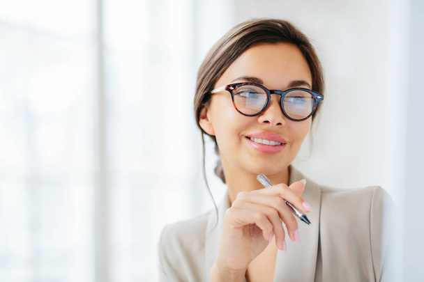 Smiling prosperous lady work in business sphere, holds pen, wears elegant clothes, has healthy skin, minimal makeup, looks somewhere, poses indoor against blurred white background, copy space left - Photo, Image