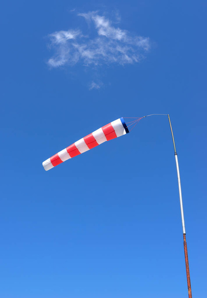 Red and white windsock made of fabric hangs almost horizontally on a pole against a blue sky with a slight cloud - 写真・画像
