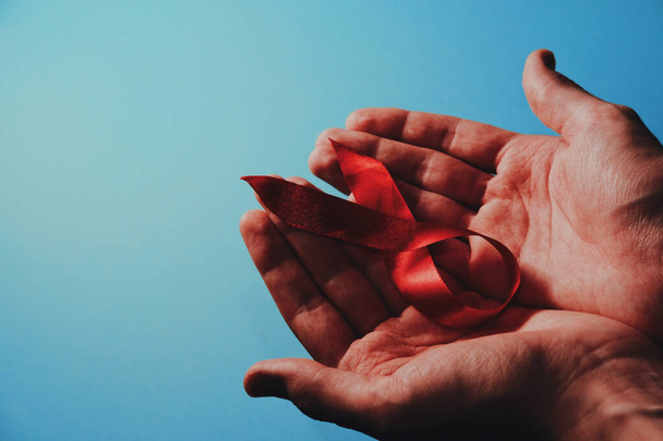 Día Mundial del SIDA en diciembre mes campaña. Primer plano de la conciencia de la cinta roja en las manos del hombre en el fondo azul. Símbolo de sensibilización y apoyo a las personas que viven con el VIH. Copiar espacio
. - Foto, Imagen