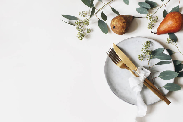Autumn thanksgiving table place setting. Golden cutlery, porcelain plate, berry eucalyptus leaves and branches, silk ribbon and pear fruit on white table background. Fall wedding. Flat lay, top view. - 写真・画像