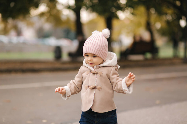 Pieni vauva kävelee puistossa 11 kuukautta. Onnellinen vauva söpö ruskea takki, vaaleanpunainen hattu ja denim farkut
 - Valokuva, kuva