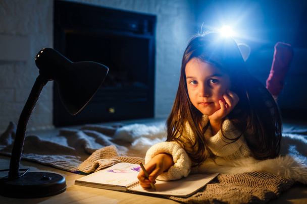ein hübsches kleines Mädchen schreibt in Notizbuch auf dem Boden unter der Lampe - Foto, Bild