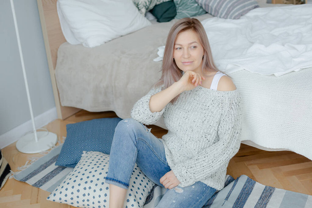 Blonde young woman in grey  pullover is near bed - 写真・画像