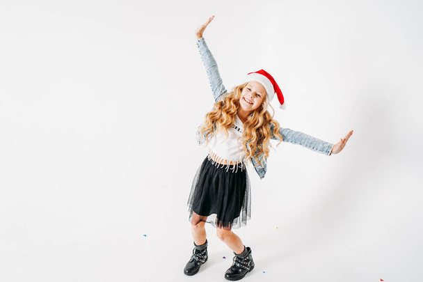 Happy fashionably dressed curly hair tween girl in santa hat, denim jacket and black tutu skirt on white background with colorful confetti - Fotografie, Obrázek