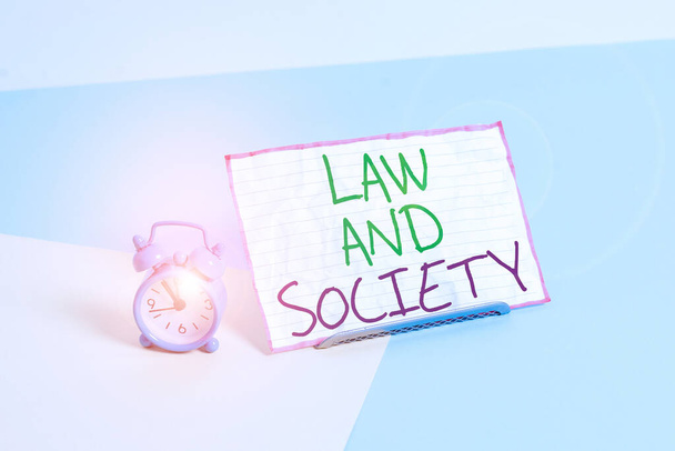 Handwriting text writing Law And Society. Concept meaning Address the mutual relationship between law and society Mini size alarm clock beside a Paper sheet placed tilted on pastel backdrop. - Photo, Image